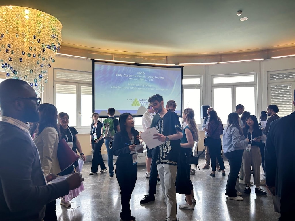 A group of people in a room engage in conversations and networking. A screen in the background displays information about an "Early Career Networking EGU Lounge" event.