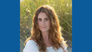 A woman with wavy brown hair wearing a white top, outdoors with greenery and sunlight in the background.
