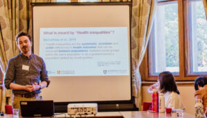 A person delivers a presentation titled "What is meant by 'Health inequalities'?" to a group of seated attendees in a room with patterned curtains and large windows.
