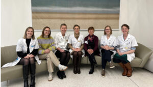 Seven healthcare professionals, dressed in white lab coats and casual attire, are seated on a couch in a hospital or clinic setting, smiling at the camera. A landscape painting hangs behind them.