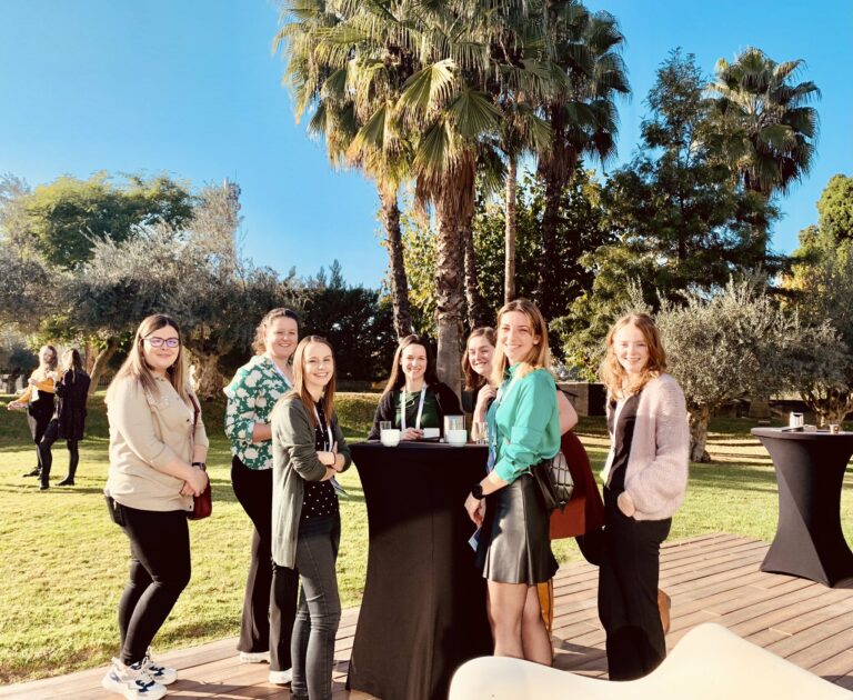 A group of people standing outdoors around a tall table, smiling, with palm trees and clear blue sky in the background.