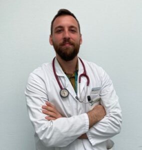 A person in a white coat with a stethoscope around their neck stands against a plain background. They have their arms crossed.