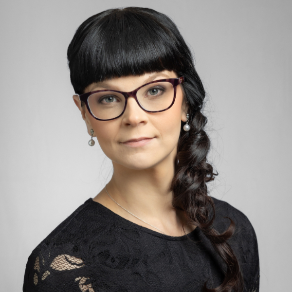 Person with long dark hair braided to the side, wearing glasses and a black lace top, against a plain background.