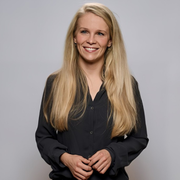 Smiling person with long blonde hair and a black shirt stands against a plain gray background.