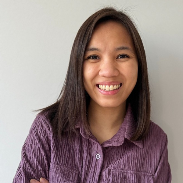A person with shoulder-length dark hair smiles while wearing a purple textured blouse, standing against a plain light-colored background.