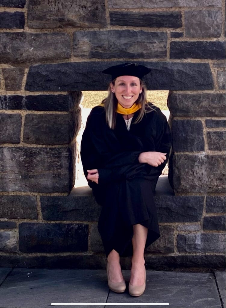 A person in a graduation cap and gown is sitting in a stone wall opening, smiling at the camera.