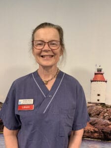 A woman in a blue medical uniform stands smiling in front of a scenic backdrop featuring a lighthouse.
