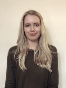 A woman with long, straight blonde hair wearing an olive green top stands in front of a plain, light-colored wall.