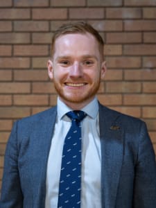 A man with short red hair and a beard, wearing a grey suit, white shirt, and blue tie, stands smiling in front of a brick wall.
