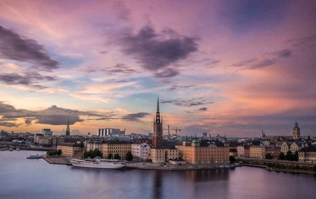 A cityscape with historic buildings and a church spire alongside a body of water at sunset, under a colorful pink and purple sky.