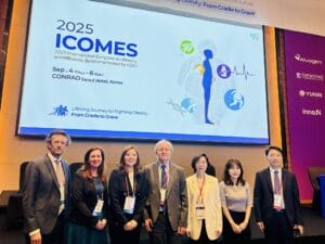 Seven professionals stand in front of a screen displaying information about the 2025 ICOMES conference on obesity and metabolic syndrome at Conrad Seoul Hotel, South Korea.