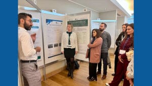A man presents research on a poster to a group of attendees at an indoor academic conference, all standing and listening attentively.