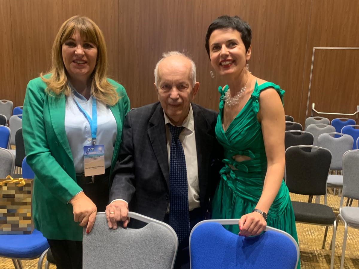 Three people pose together in a conference room with empty chairs; two women stand on either side of an older man seated in the center.