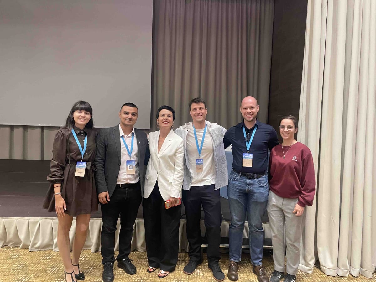 Six people stand side by side indoors, wearing conference name badges, smiling and posing in front of a stage with curtains.