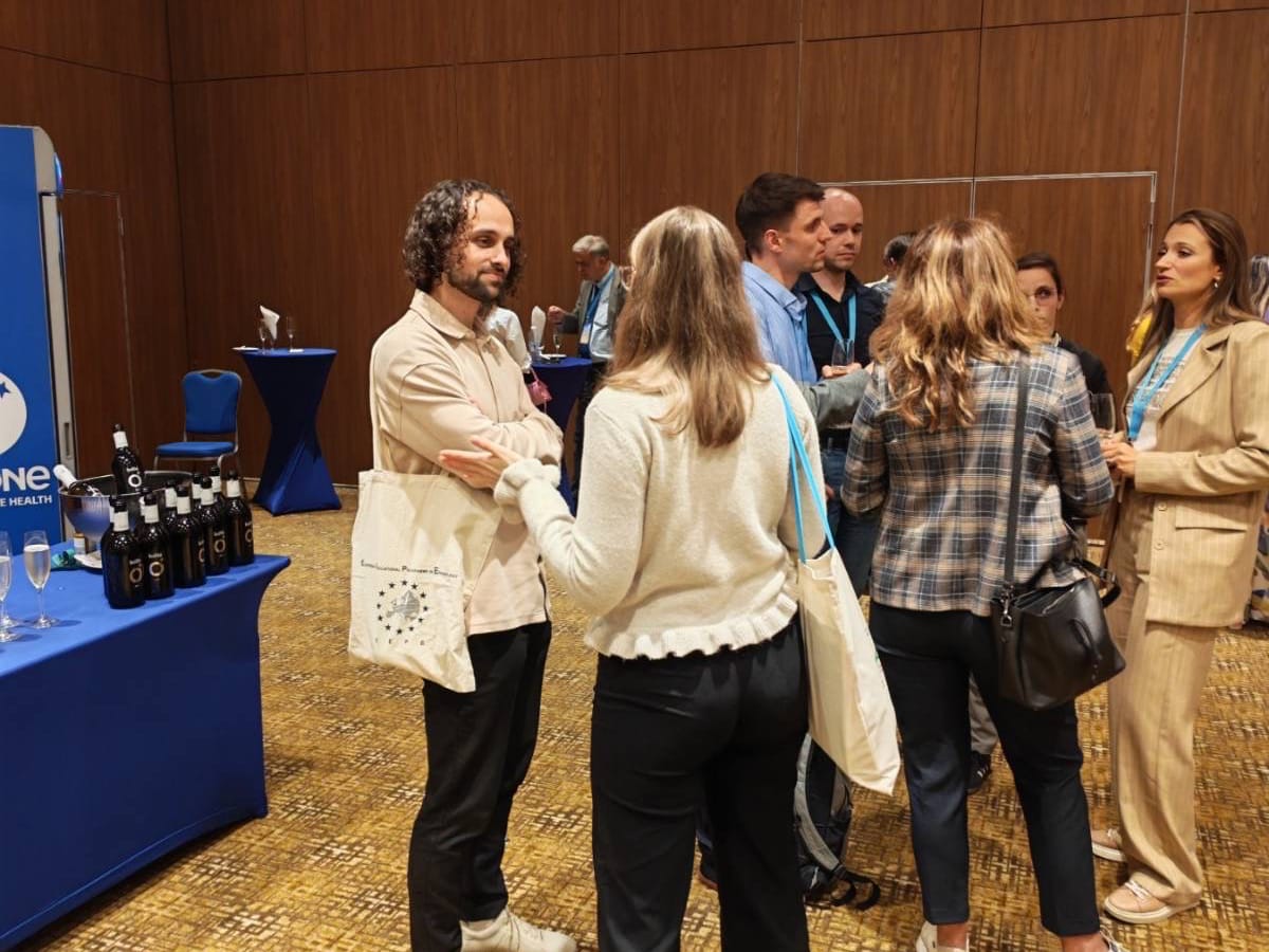 A group of people stand and converse in a conference room with brown walls and carpeted floors. Bottles and glasses are on a nearby table. Most people wear conference lanyards.