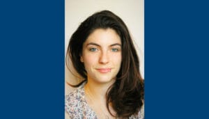 A woman with long dark hair and light eyes, wearing a patterned blouse and a lip piercing, poses for a headshot against a plain background.
