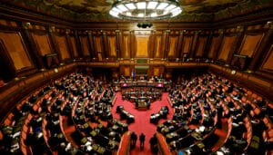 A wide view of a parliamentary chamber with rows of desks and officials seated, observing proceedings under a large ornate ceiling light.