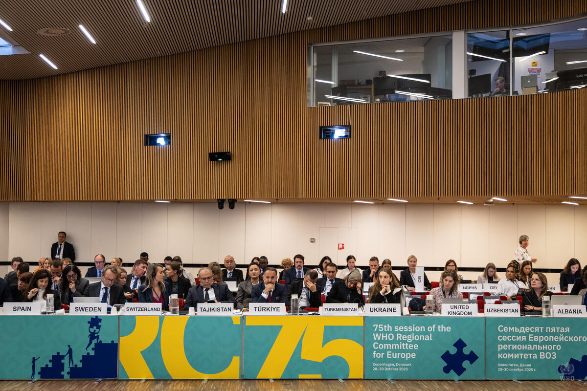 Delegates from various countries seated at tables during the 75th session of the WHO Regional Committee for Europe, with country nameplates displayed.