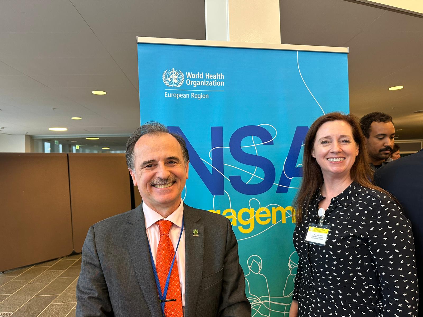 Two people stand smiling in front of a World Health Organization European Region banner indoors at what appears to be a professional event or conference.
