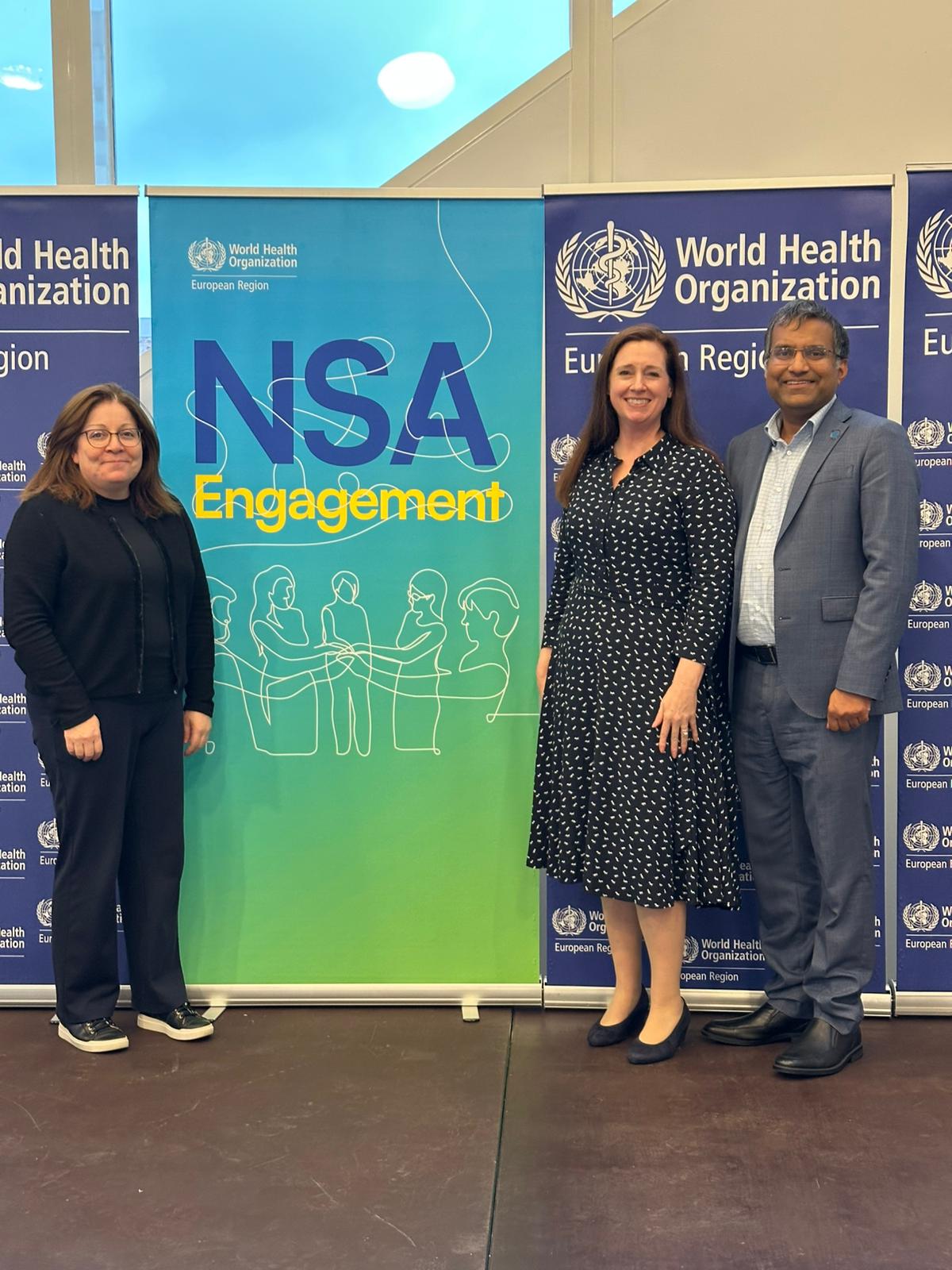 Three people stand in front of a banner reading "NSA Engagement" and World Health Organization signs, posing for a photo indoors.