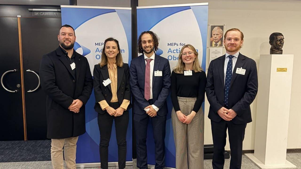Five people stand side by side in business attire, posing in front of banners that read "MEPs for Action on Obesity" in an indoor setting with two busts visible.