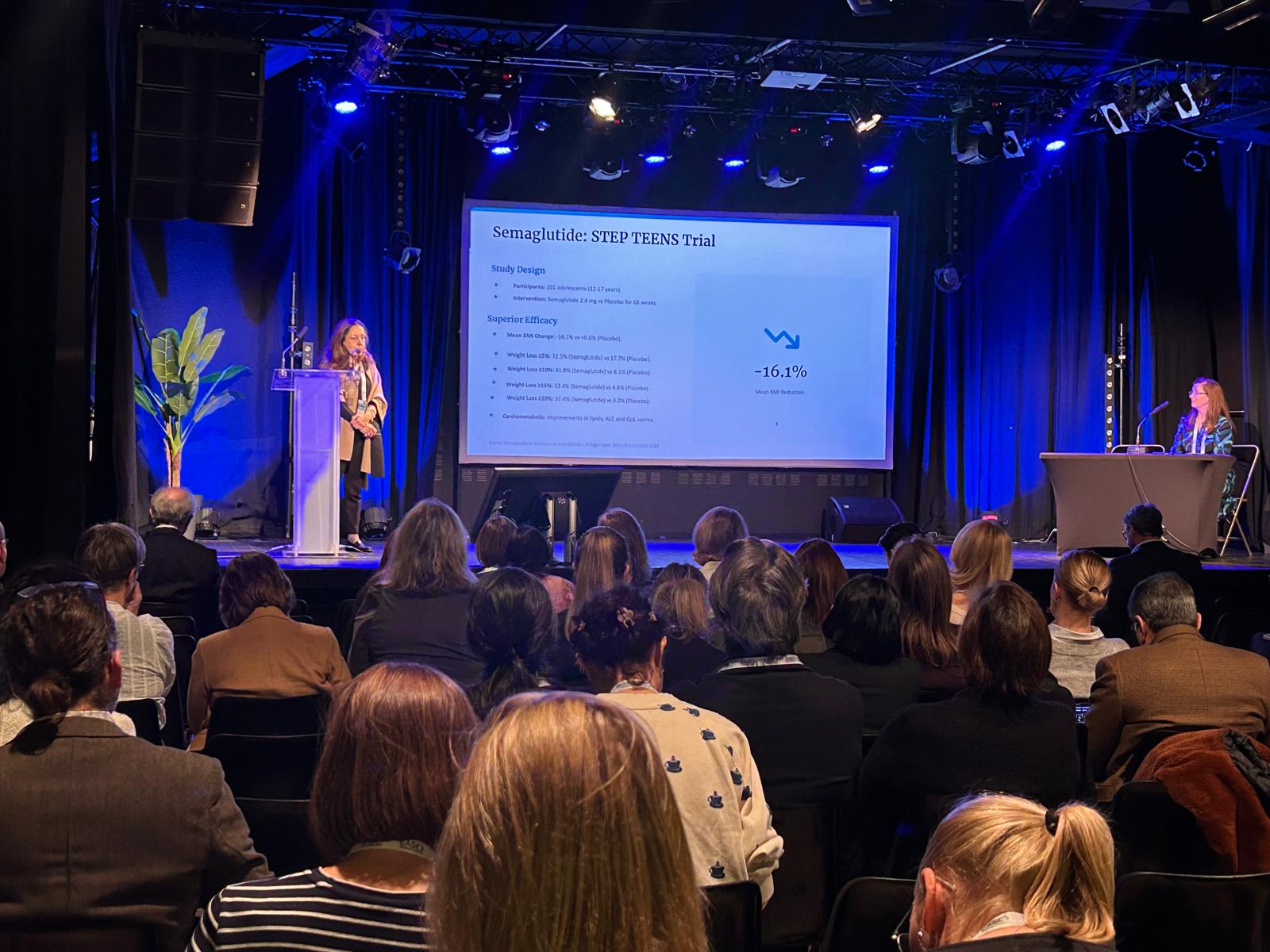 A speaker presents slides about the Semaglutide STEP TEENS Trial to an audience in a dimly lit conference room, while another person sits at a table on stage.