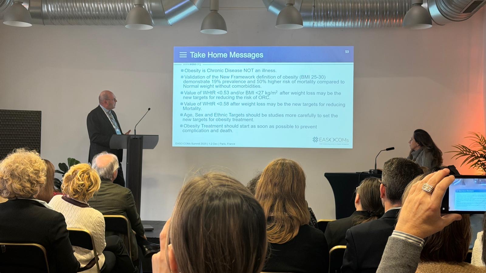 A speaker presents a slide titled "Take Home Messages" on obesity to an audience in a conference room, with attendees listening and one person recording the event on a phone.