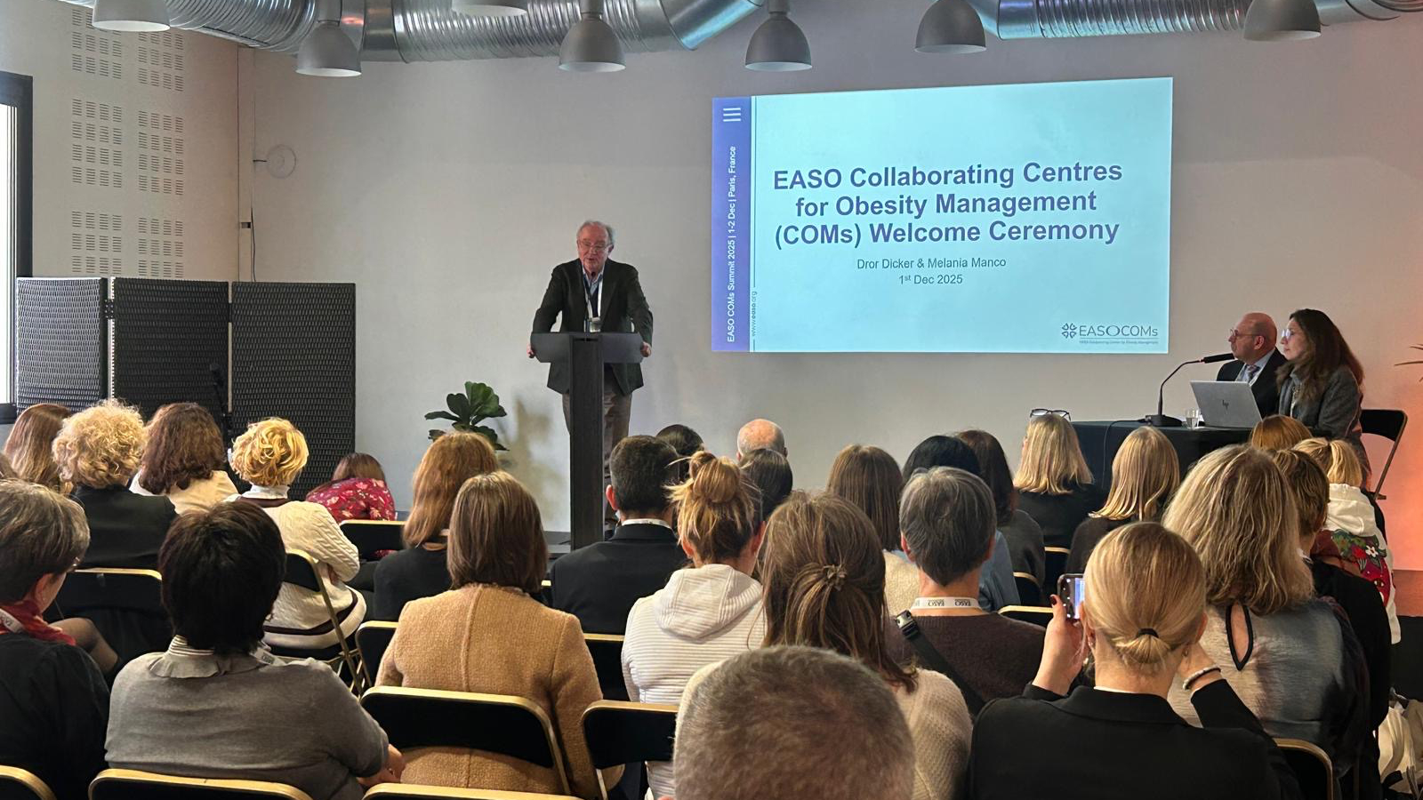 A speaker addresses an audience at the EASO Collaborating Centres for Obesity Management (COMs) Welcome Ceremony. A presentation slide is displayed behind the speaker.
