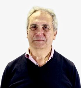 An older man with gray hair and a slight smile is standing against a plain white background, wearing a dark sweater over a light pink collared shirt.