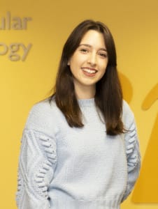 A woman with straight brown hair wearing a light blue jumper stands and smiles in front of a yellow wall with partial text visible.