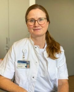 A woman wearing glasses and a white medical coat with an ID badge sits indoors, facing the camera and smiling slightly.