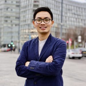 A man wearing glasses and a blue blazer stands outdoors with arms crossed, smiling, in front of a modern office building and parked cars.