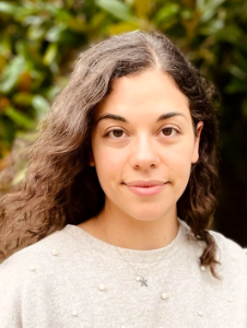A woman with long, wavy brown hair and a light grey jumper stands outdoors in front of green foliage, looking at the camera with a neutral expression.