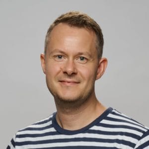 A man with short light brown hair and a trimmed beard, wearing a navy and white striped shirt, poses against a plain light grey background.