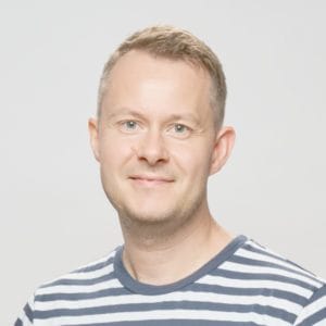 A man with short blond hair and a light beard, wearing a striped shirt, poses in front of a plain light background.
