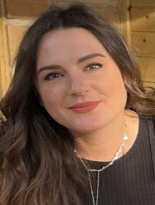 A woman with long brown hair, wearing a black top and layered necklaces, is sitting in front of a wooden background, looking slightly to the side with a neutral expression.