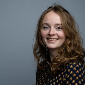A young woman with wavy brown hair smiles at the camera, standing in front of a plain grey background. She is wearing a dark shirt with a yellow pattern.