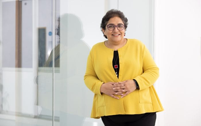 A woman wearing glasses and a yellow jacket stands indoors, smiling, with her hands clasped in front of her near a glass wall.