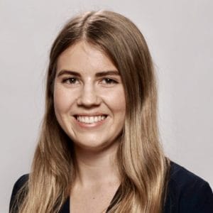 A woman with long, straight, light brown hair is smiling at the camera against a plain, light-coloured background. She is wearing a dark top.