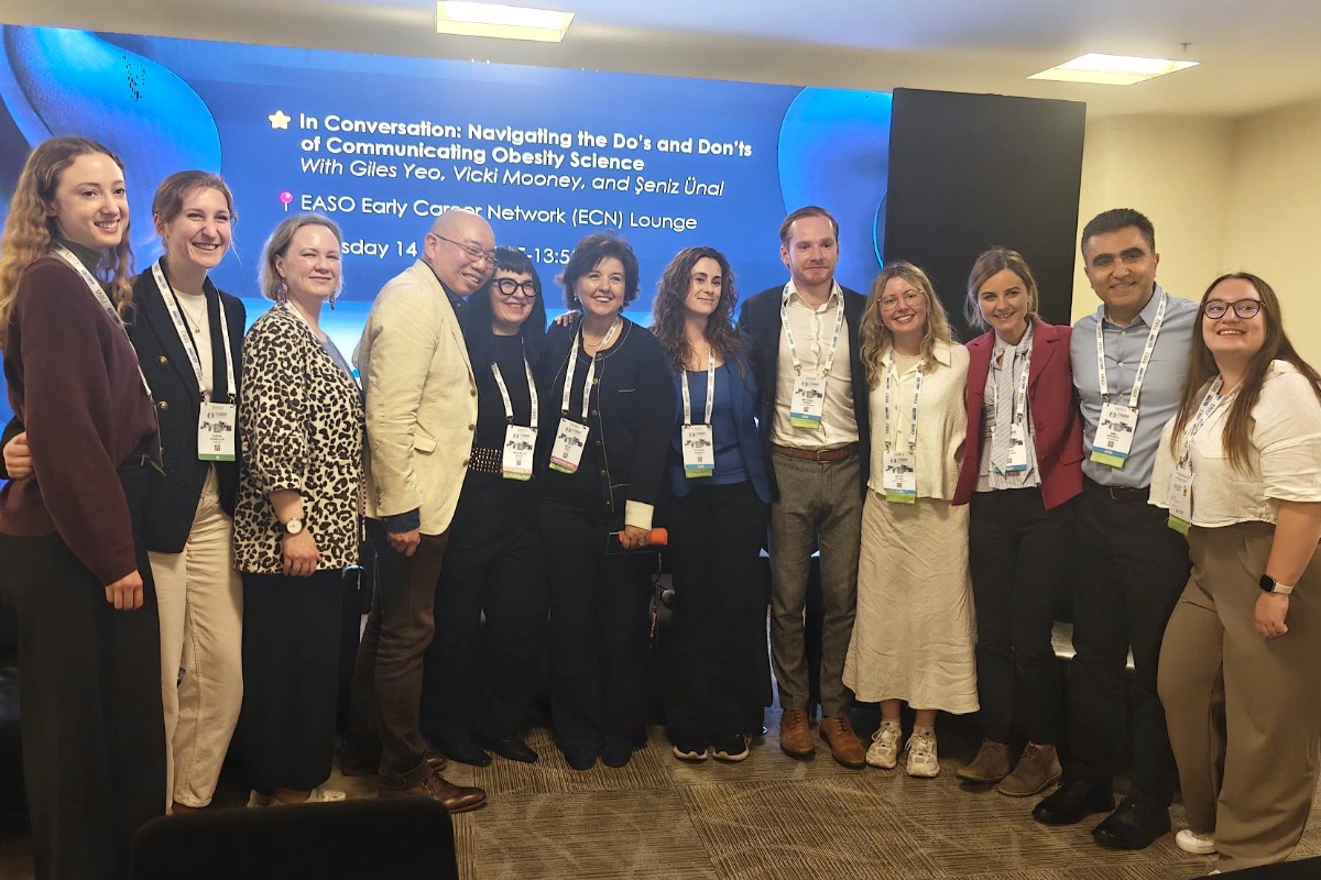 Thirteen people stand in a row indoors, posing and smiling for a group photo in front of a presentation screen about communicating obesity science.