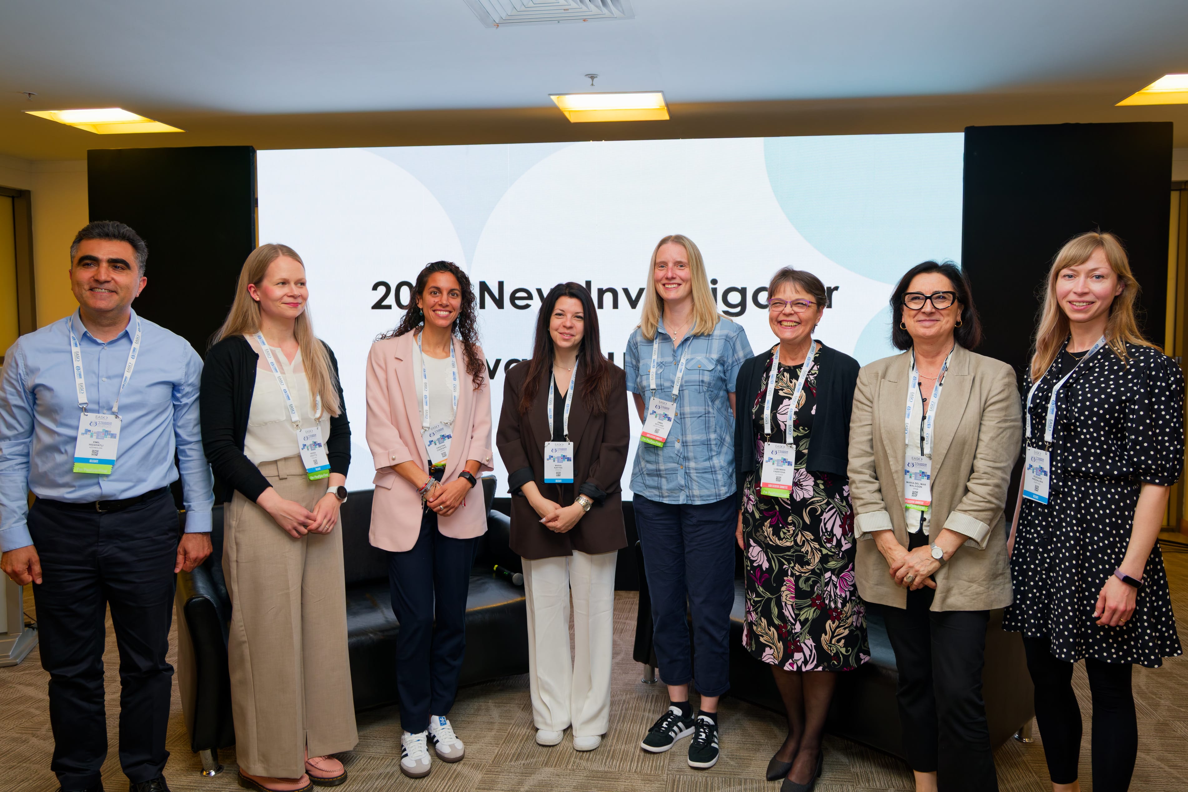 Eight people stand in a row, wearing business or casual attire, posing for a group photo at a professional event with conference badges visible.
