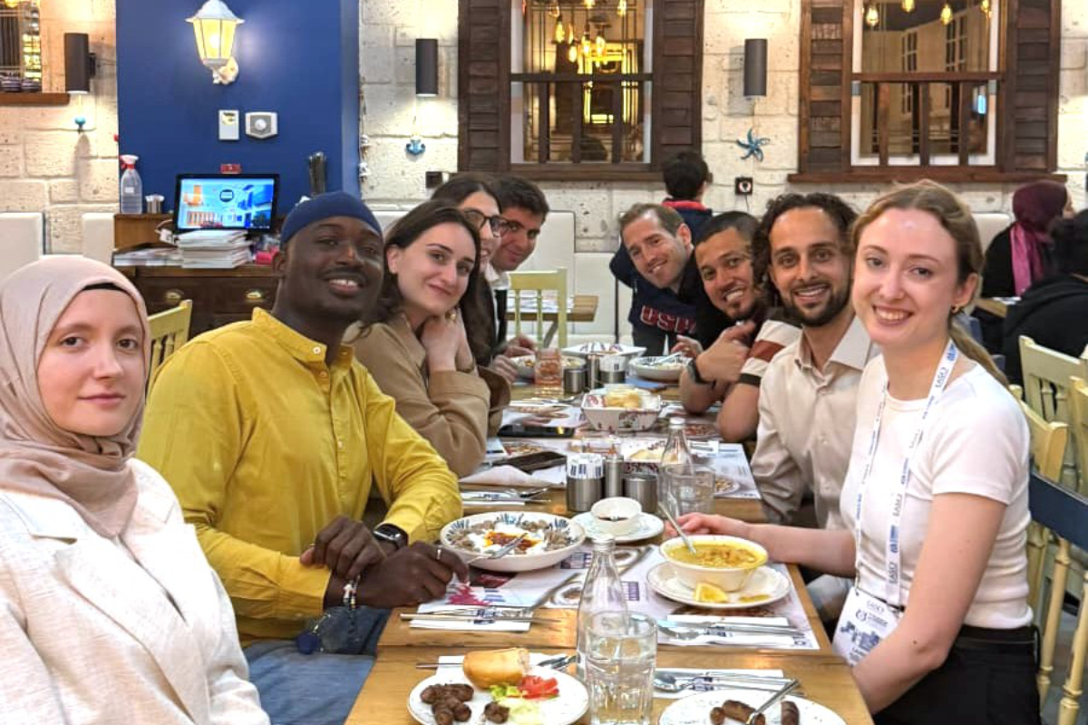 A group of eight people sit around a restaurant table, smiling at the camera with finished and half-eaten dishes in front of them.