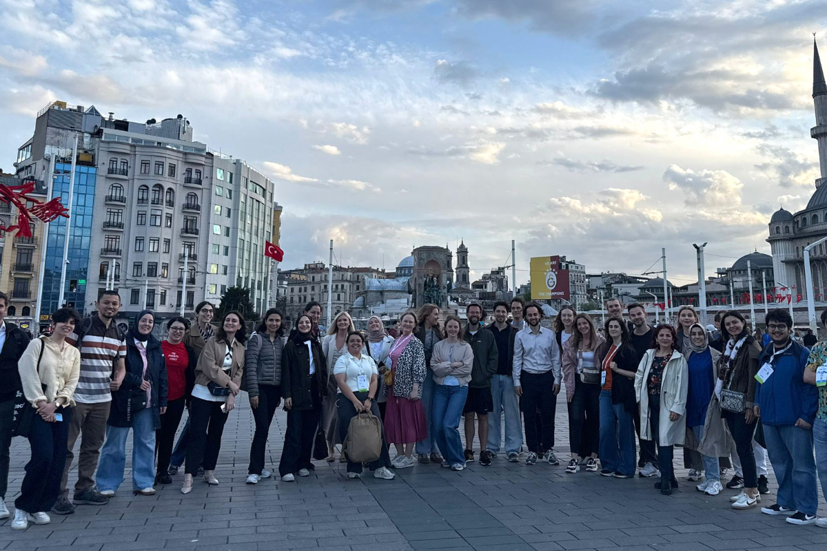 A large group of people pose together outdoors in an urban square with buildings, flags, and a mosque visible in the background under a partly cloudy sky.