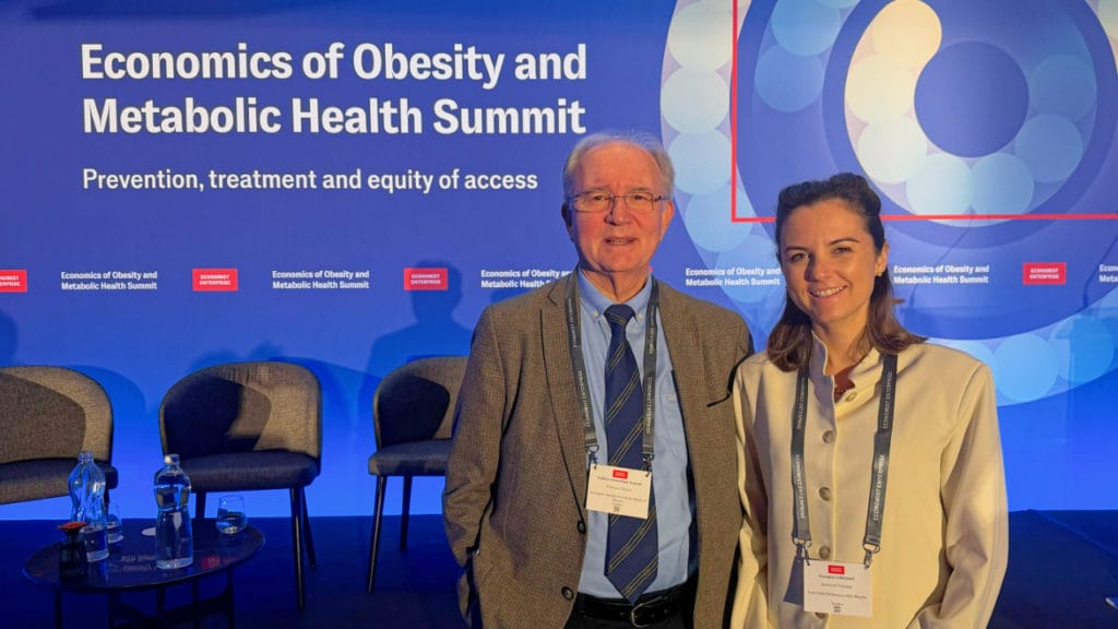 Two conference attendees stand and smile in front of a blue backdrop at the Economics of Obesity and Metabolic Health Summit. Three empty chairs and a table are visible beside them.