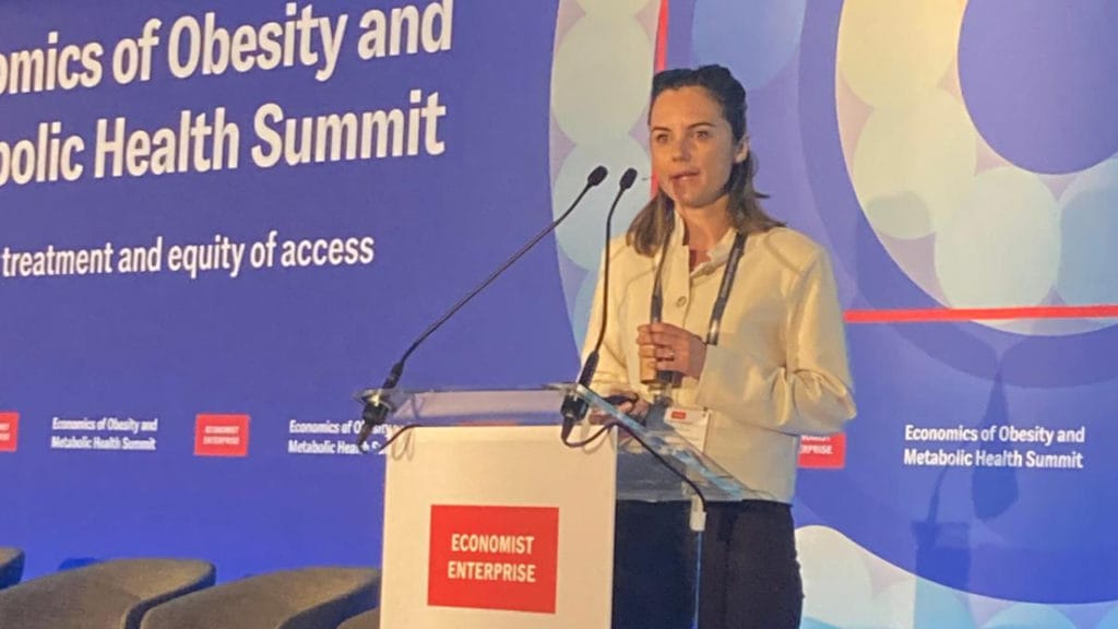 A woman speaks at a lectern during the Economics of Obesity and Metabolic Health Summit, with event banners in the background.