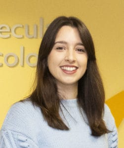 A young woman with long brown hair, wearing a light blue jumper, smiles at the camera against a yellow background with partial text visible.