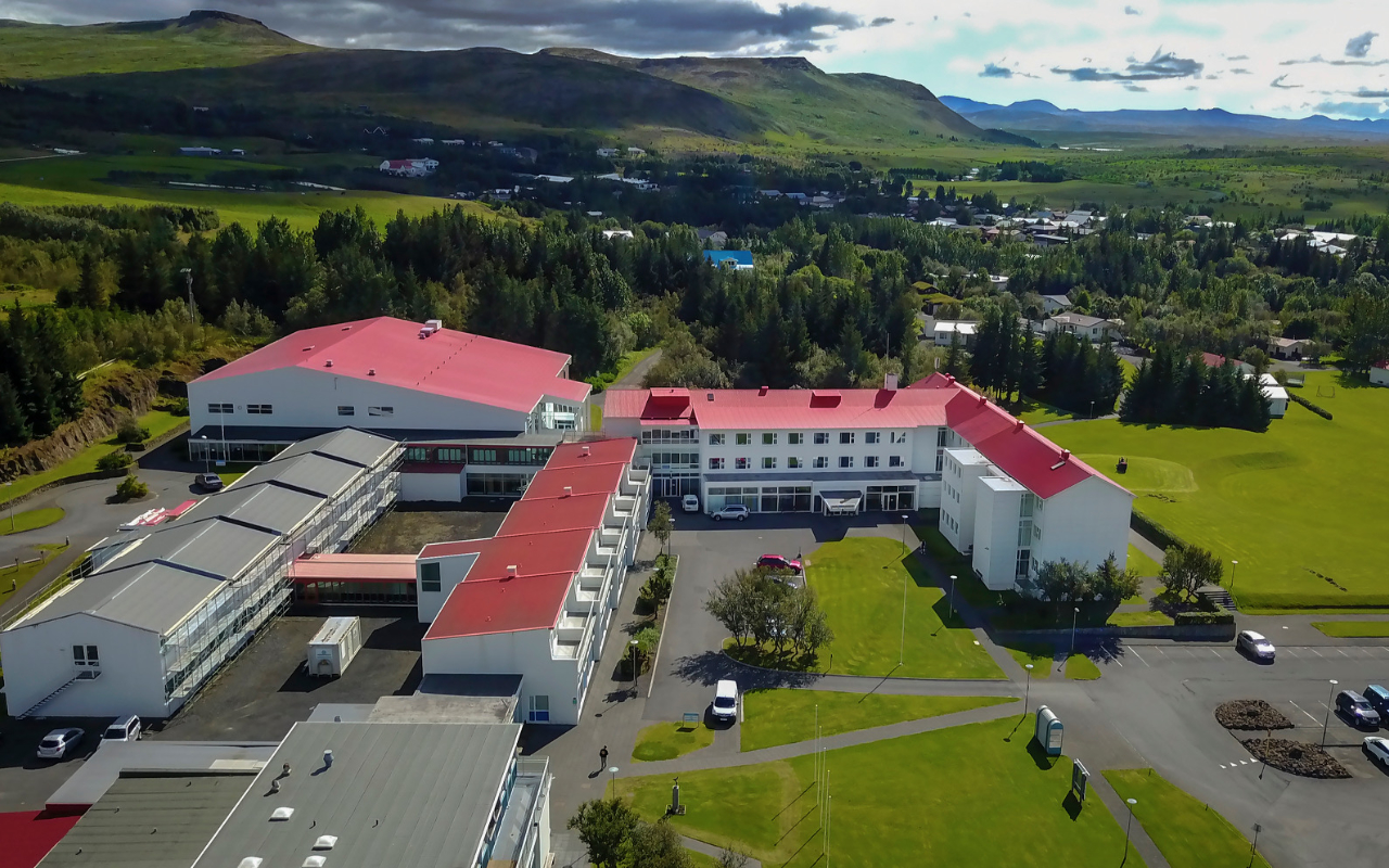 Aerial view of a school campus with red-roofed buildings surrounded by greenery, trees, and distant mountains under a cloudy sky.