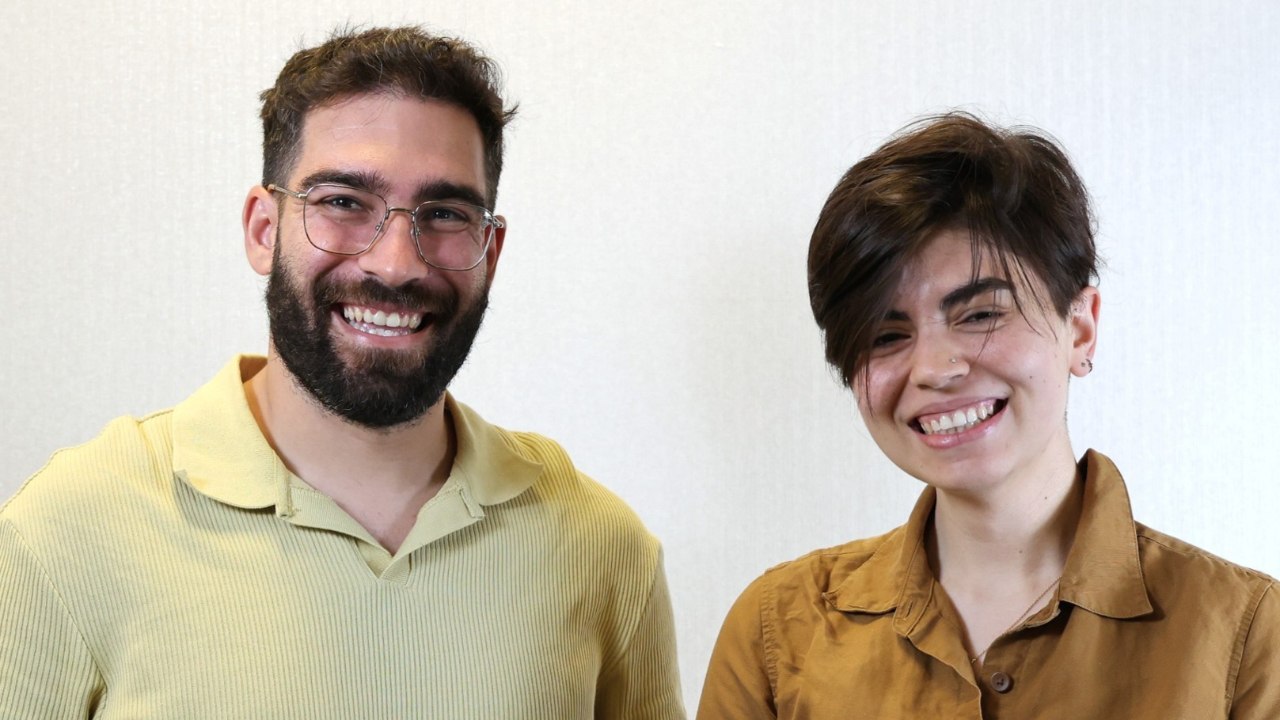 Two people standing side by side, both smiling at the camera. The person on the left has glasses and a beard; the person on the right has short hair. Both wear light-coloured shirts.