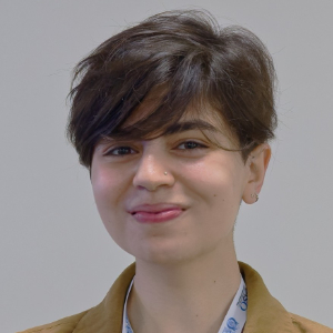 A person with short, dark hair is smiling slightly, wearing a brown jacket and a collared shirt, against a plain light background.