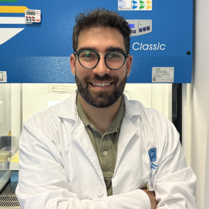A man in a white lab coat stands smiling in front of laboratory equipment with his arms folded.